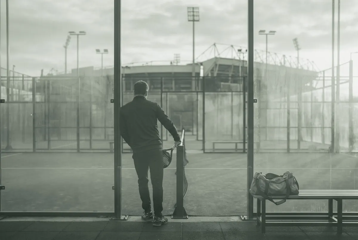 Sporter met racket bij een padelbaan in de schemering, op de achtergrond vaag de lichtmasten van een voetbalstadion